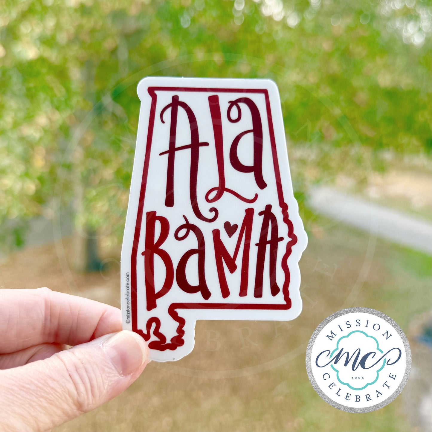 Hand holding a 'Alabama' shaped sticker with blurred greenery in the background