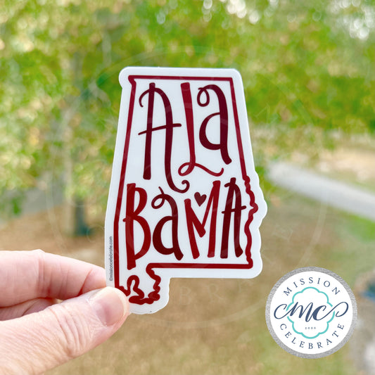 Hand holding a 'Alabama' shaped sticker with blurred greenery in the background